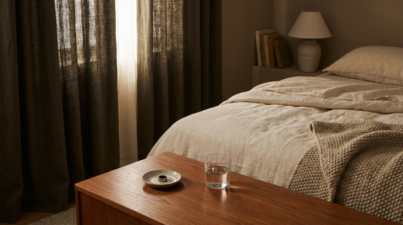 A dim, tidy bedroom at dusk with a neatly made bed, a sleep-tracking ring and water glass on the nightstand, and blackout curtains letting in a thin line of light.